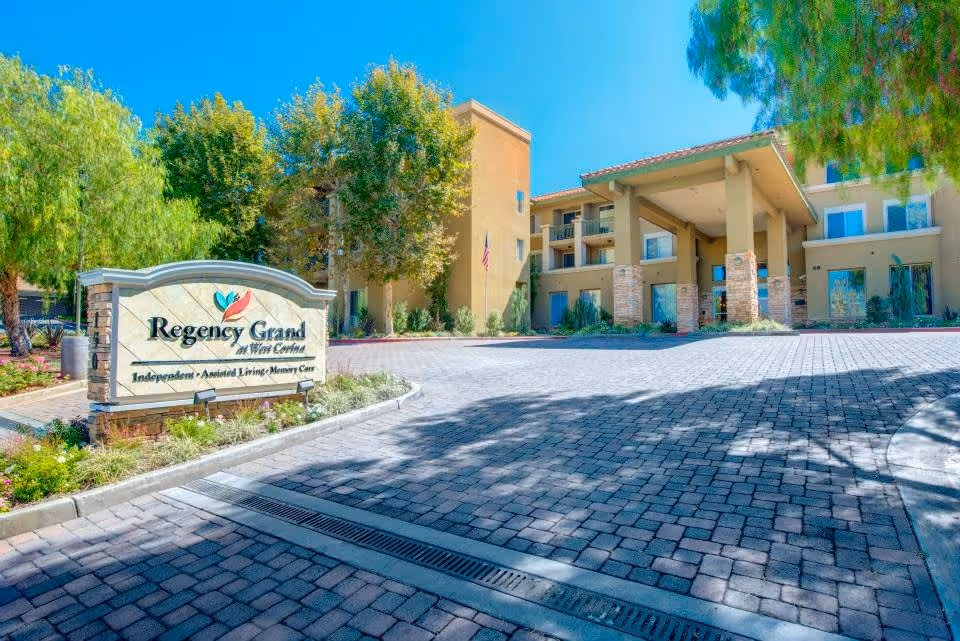 Exterior view of Regency Grand at West Covina senior living facility with a paved driveway, landscaped greenery, and a large entrance canopy under a clear blue sky.