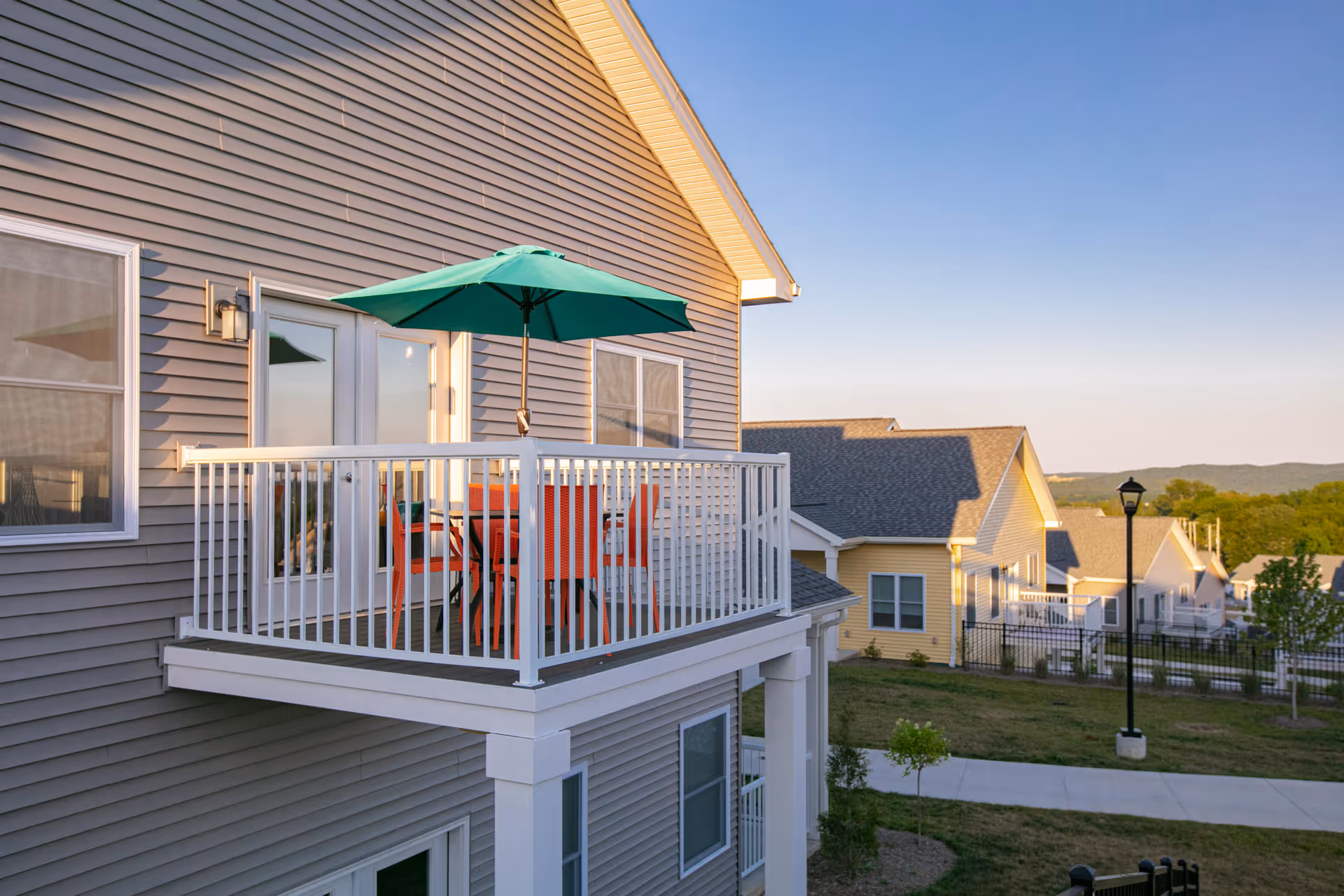 A balcony attached to a residential building with a white railing, a green patio umbrella, and orange chairs around a table. The building has gray siding and white trim. In the background, there are other similar residential buildings, a streetlamp, and a clear sky during sunset.