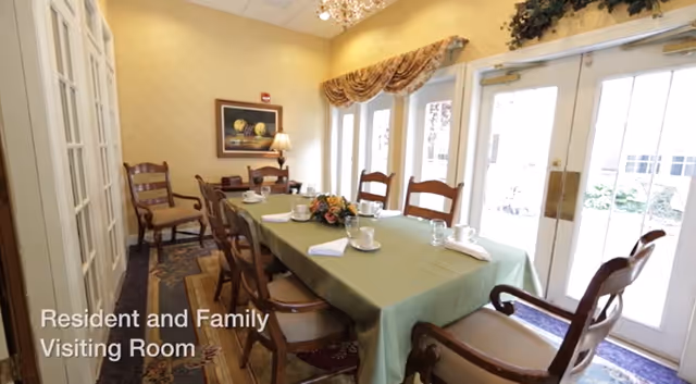 A dining room with a long table covered with a green tablecloth, set with cups, saucers, and napkins. The room has wooden chairs around the table, a painting on the wall, a lamp, and large glass doors letting in natural light. A floral centerpiece is on the table.