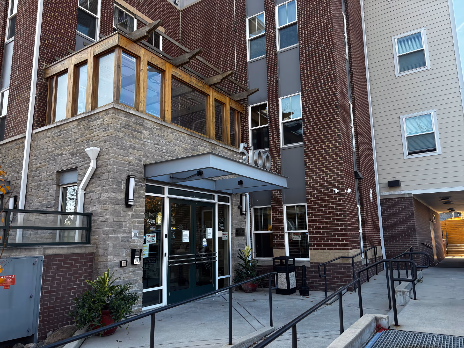 Entrance of a senior living facility with a stone and brick exterior, glass double doors, a metal awning above the entrance, and a ramp with handrails leading up to the door. There are potted plants near the entrance and multiple windows on the building.