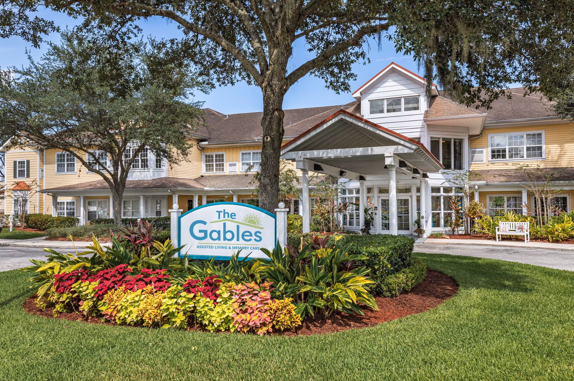 Exterior view of The Gables of Jacksonville assisted living and memory care facility, showing a two-story yellow building with white trim, a covered entrance, well-maintained landscaping with colorful flowers and green grass, and a sign in front displaying the facility's name.