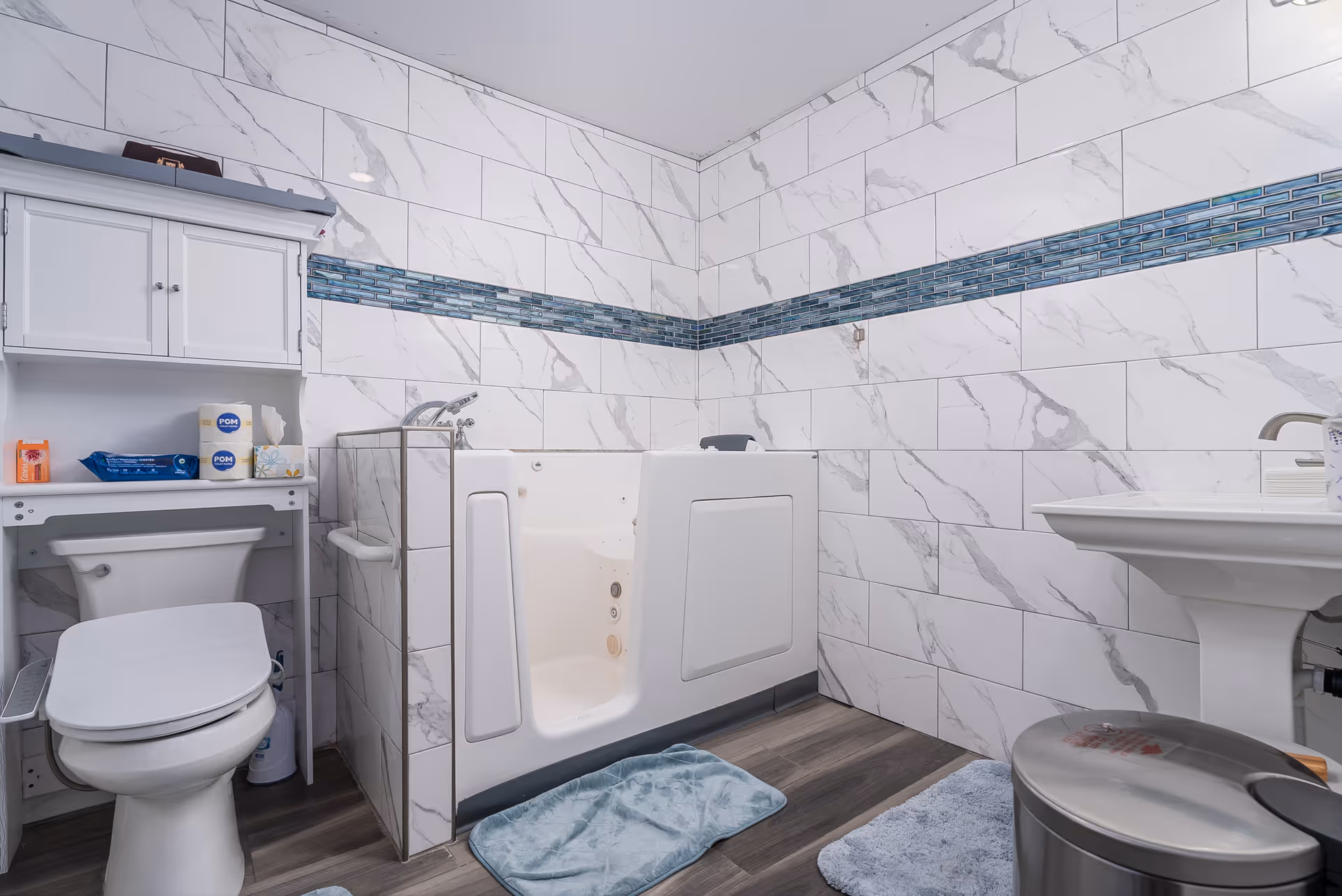 A clean and modern bathroom with white marble-patterned tiles on the walls and a horizontal strip of blue mosaic tiles. The room features a walk-in bathtub with a door, a white toilet with a small cabinet above it holding toiletries, a white pedestal sink, and two blue bath mats on the wooden floor. A stainless steel trash can is also visible near the sink.