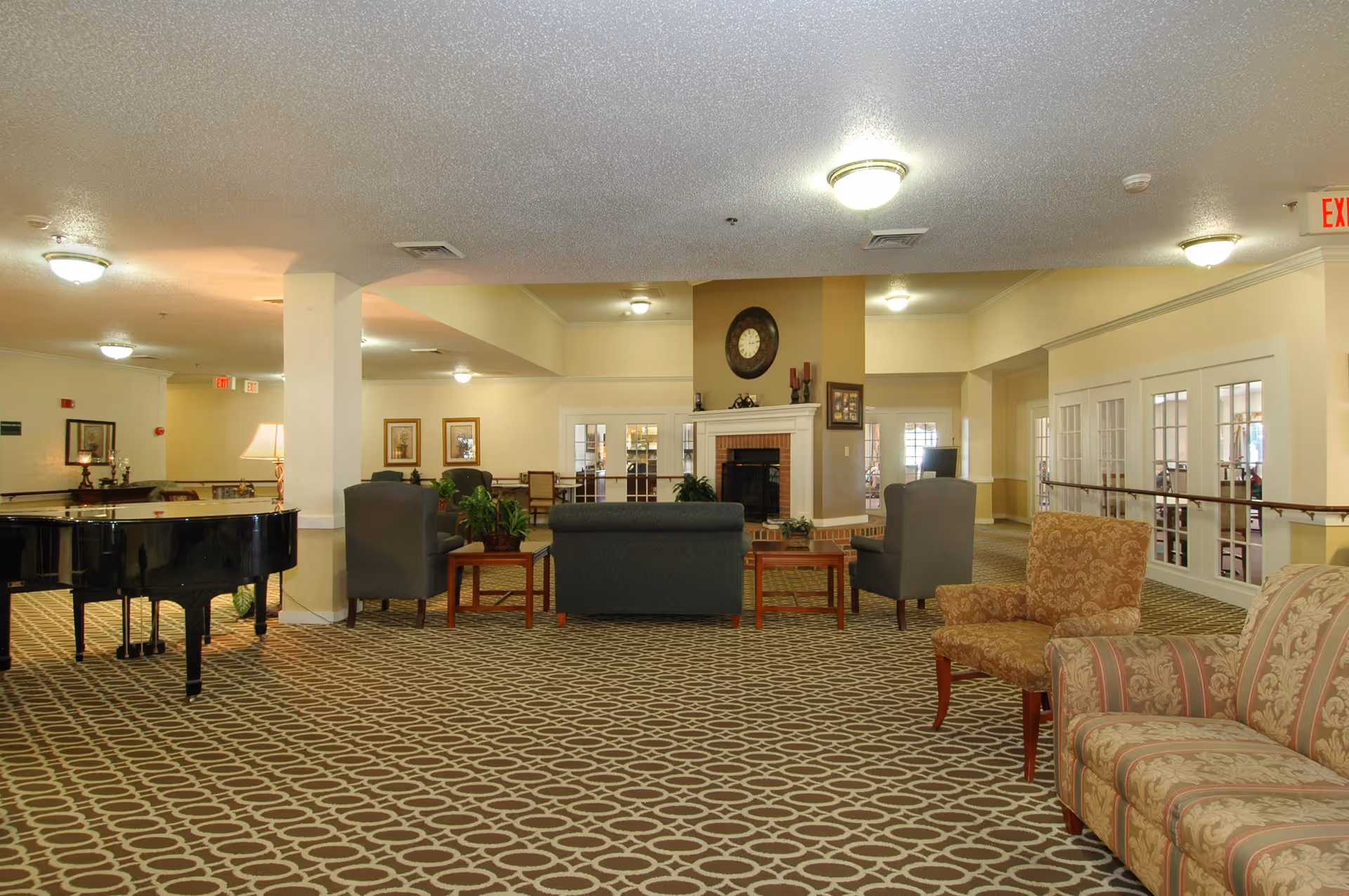 A spacious senior living facility common area with patterned carpet, multiple upholstered chairs and sofas arranged around wooden coffee tables, a black grand piano on the left, and a fireplace with a large clock above it in the center background. The room is well-lit with ceiling lights and has light-colored walls with framed artwork. French doors and an exit sign are visible in the background.