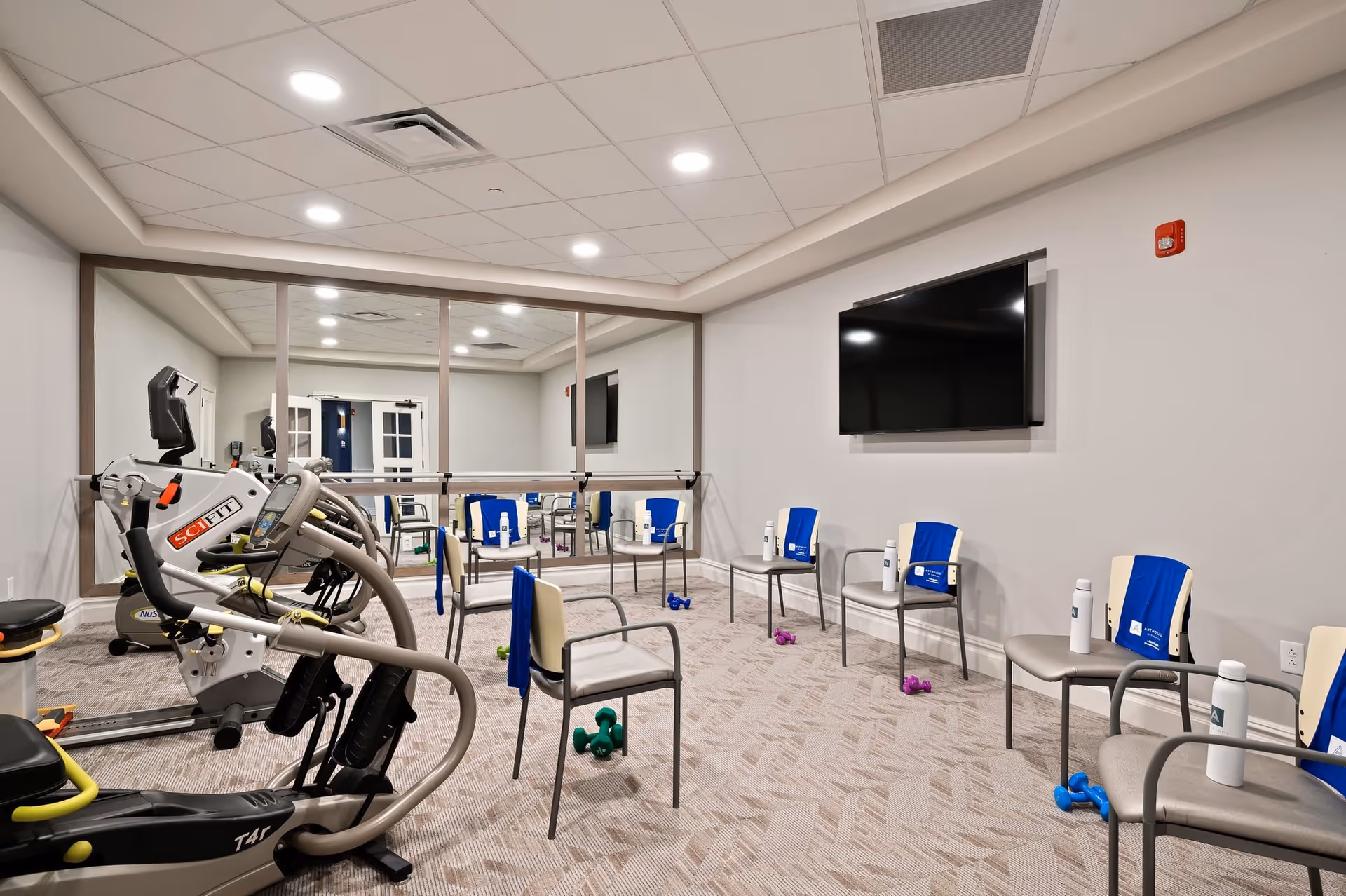 A fitness room with exercise equipment including a recumbent bike and elliptical machine. Several chairs are arranged in a semicircle, each with a blue towel and a white water bottle on or near it. Small dumbbells are placed on the carpeted floor near the chairs. A large flat-screen TV is mounted on the wall, and a large mirror covers one side of the room.