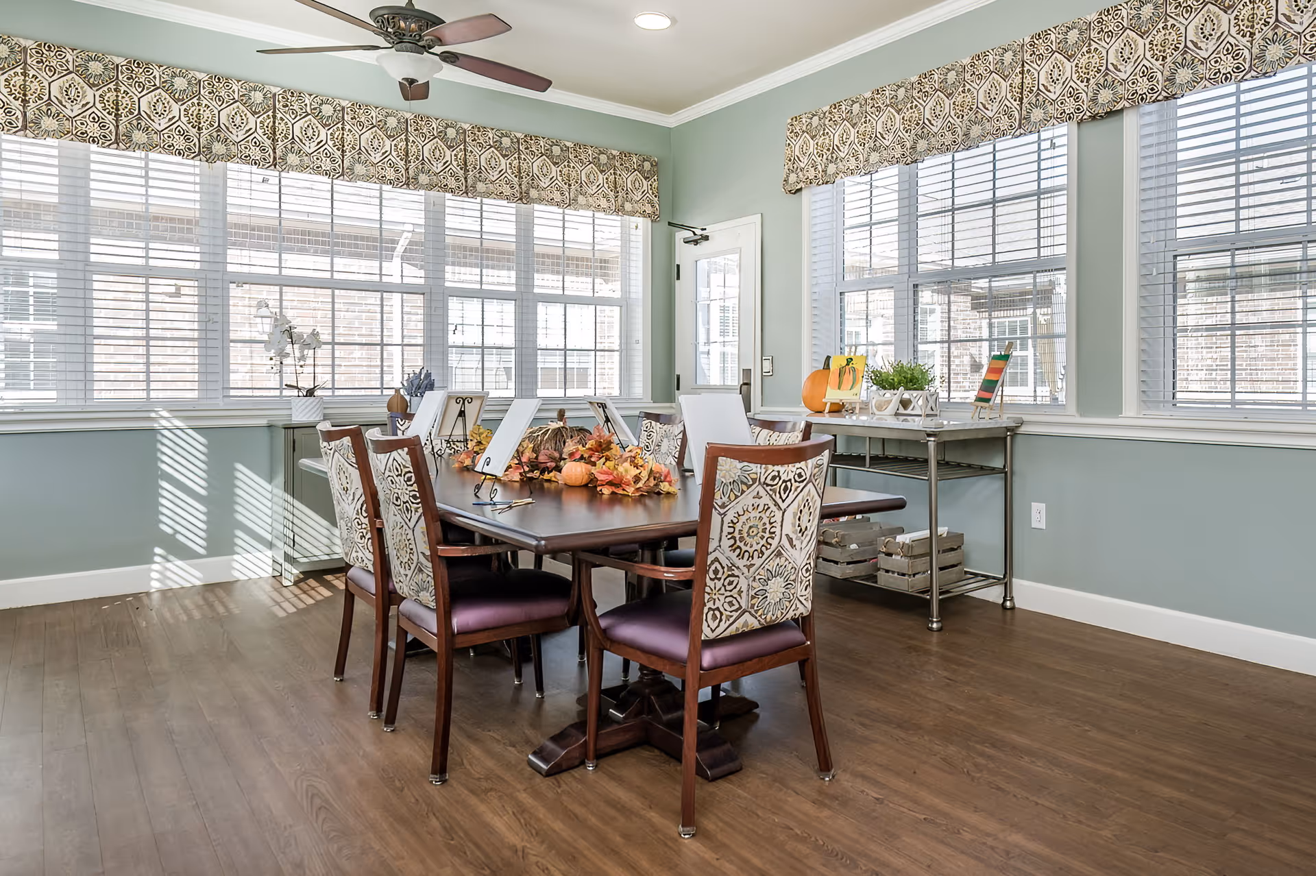 A bright dining room with a wooden table surrounded by six chairs with patterned upholstery. The table is decorated with autumn-themed centerpieces including leaves and small pumpkins. Large windows with white blinds and patterned valances let in natural light. A ceiling fan is mounted above the table, and a metal cart with decorative items is against the wall.