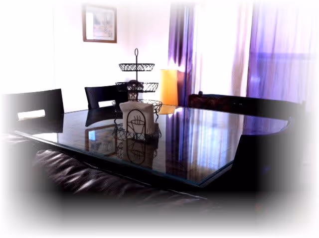 A dining room with a glass-top table surrounded by dark chairs. On the table, there is a tiered metal basket and a napkin holder. In the background, there are purple curtains and a framed picture on the wall.