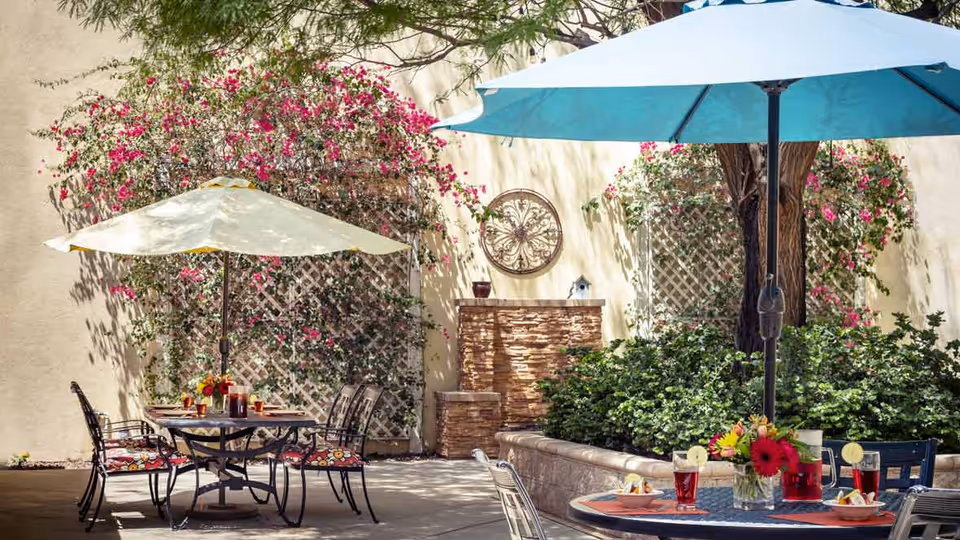 Outdoor patio area with two tables, each shaded by large umbrellas. One umbrella is light yellow and the other is light blue. The tables have chairs with floral cushions and are set with drinks and flowers. The background features a beige wall with climbing plants and a decorative circular wall ornament.