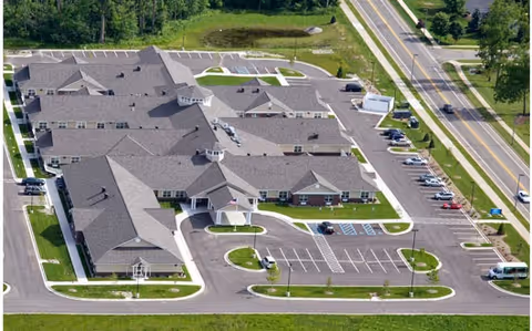 Aerial view of Cedar Creek Health Campus showing a large single-story building with multiple roof sections, surrounded by parking lots and green landscaped areas. The facility is adjacent to a road with vehicles parked along the side.