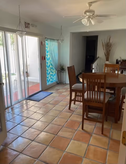 Sunlit dining area with a round wooden table and chairs on terracotta tile flooring, sliding glass doors to a patio, and a ceiling fan.