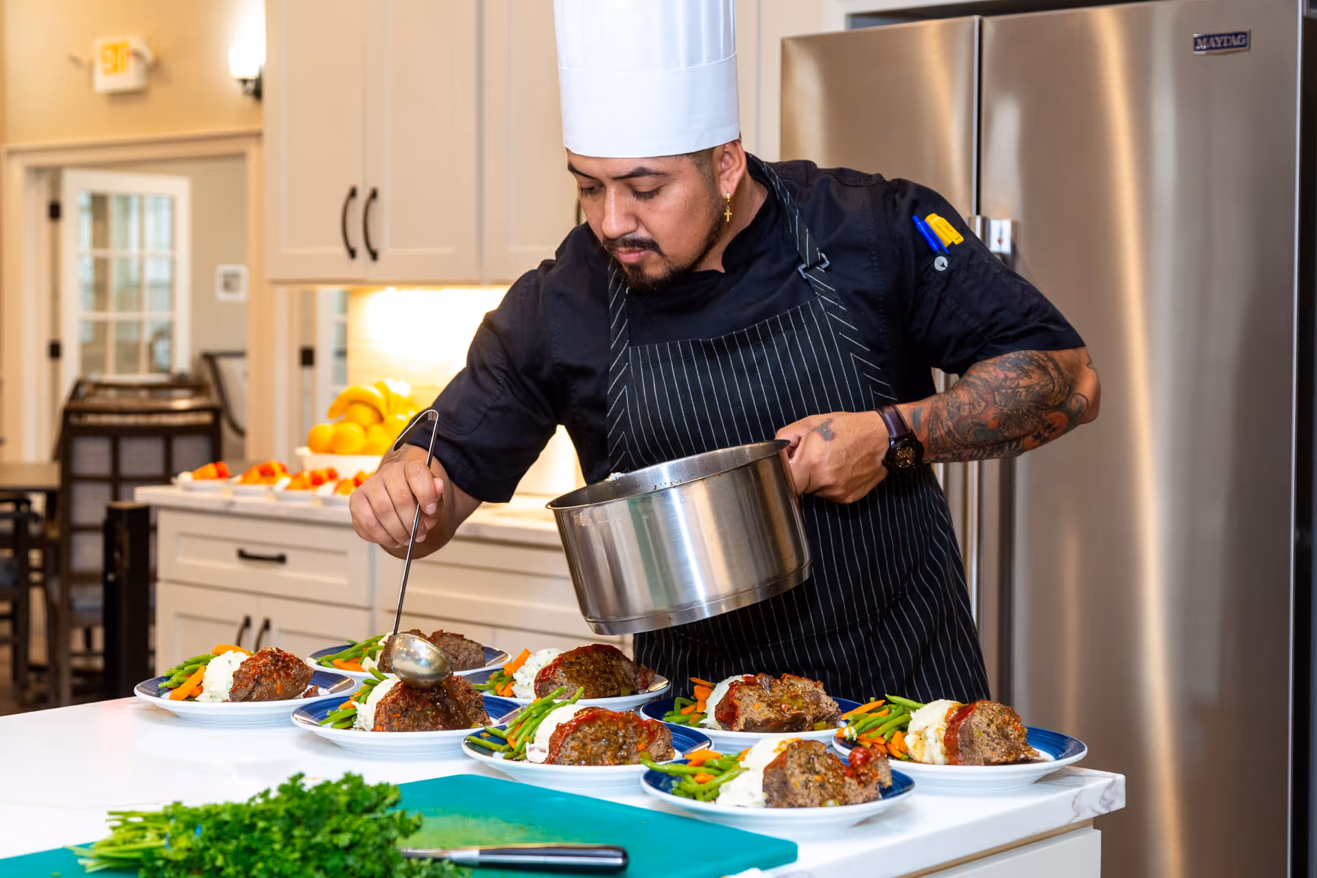 A chef in a white hat and striped apron plates multiple meals on a kitchen island.