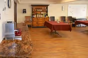 A spacious room with wooden flooring featuring a long table covered with a red tablecloth in the center, several chairs arranged along the walls, a wooden cabinet with shelves holding various items, a whiteboard on the wall, and a countertop with a coffee maker and other appliances on the left side.