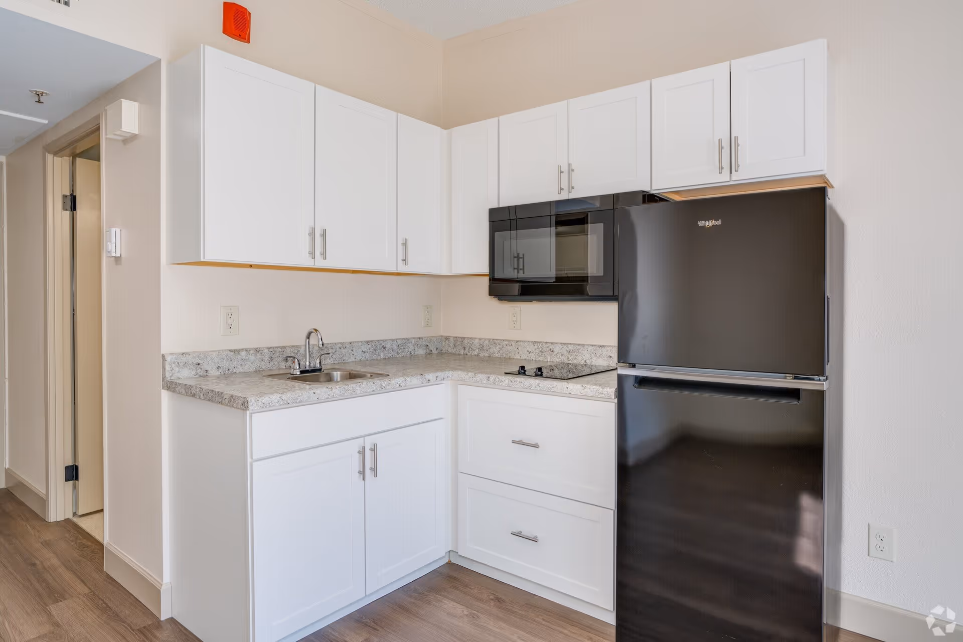 A small kitchen area with white cabinets, a granite countertop, a stainless steel sink, a black microwave mounted above a black electric stovetop, and a black refrigerator. The floor is wood, and there is a doorway visible to the left.
