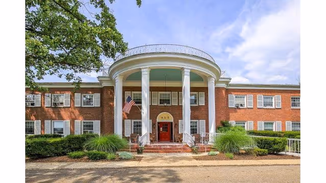 Front exterior view of a two-story brick building with white columns and a red door, surrounded by greenery and an American flag hanging near the entrance under a partly cloudy sky.