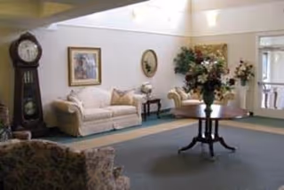 Bright common-area living room with sofas, a round table holding a floral centerpiece, a grandfather clock, and framed artwork on the walls.