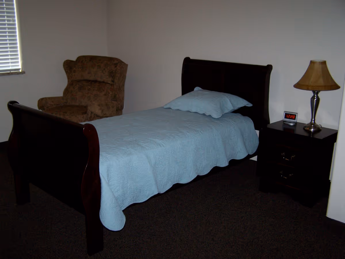 A simple bedroom with a single dark wooden bed covered with a light blue quilt and matching pillow. Next to the bed is a dark wooden nightstand with two drawers, a digital clock displaying 12:28, and a lamp with a beige lampshade. In the corner near a window with closed blinds, there is a brown upholstered armchair.