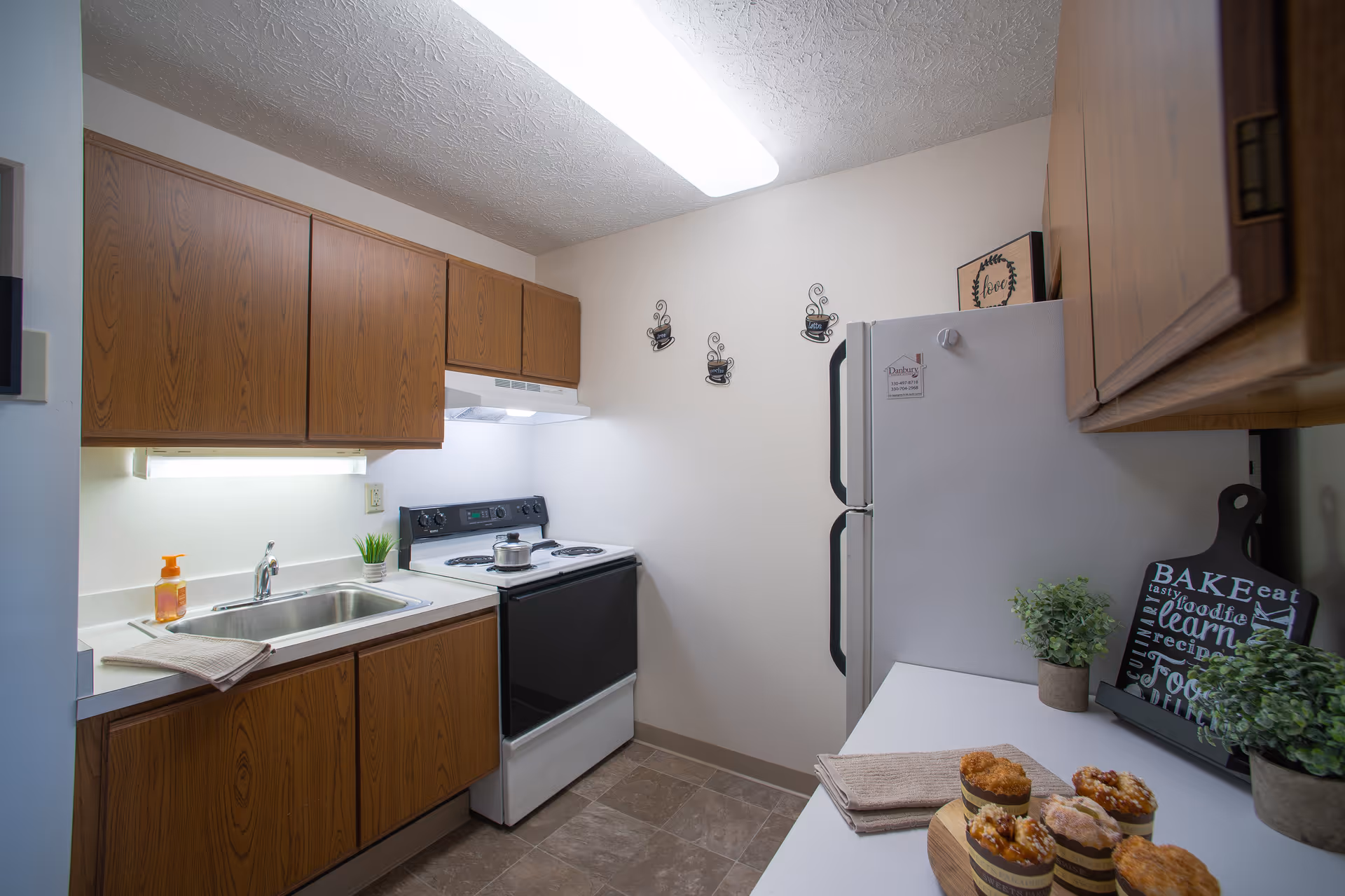 A small kitchen with wooden cabinets, a white countertop, a stainless steel sink with a soap dispenser and towel, an electric stove with a pot on it, a white refrigerator with a Danbury North Canton magnet, and a countertop with muffins, potted plants, and a decorative cutting board with food-related words.
