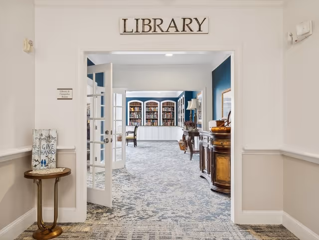 Entrance to a cozy library with a 'LIBRARY' sign above the doorway, open French doors, bookshelves and seating visible inside.