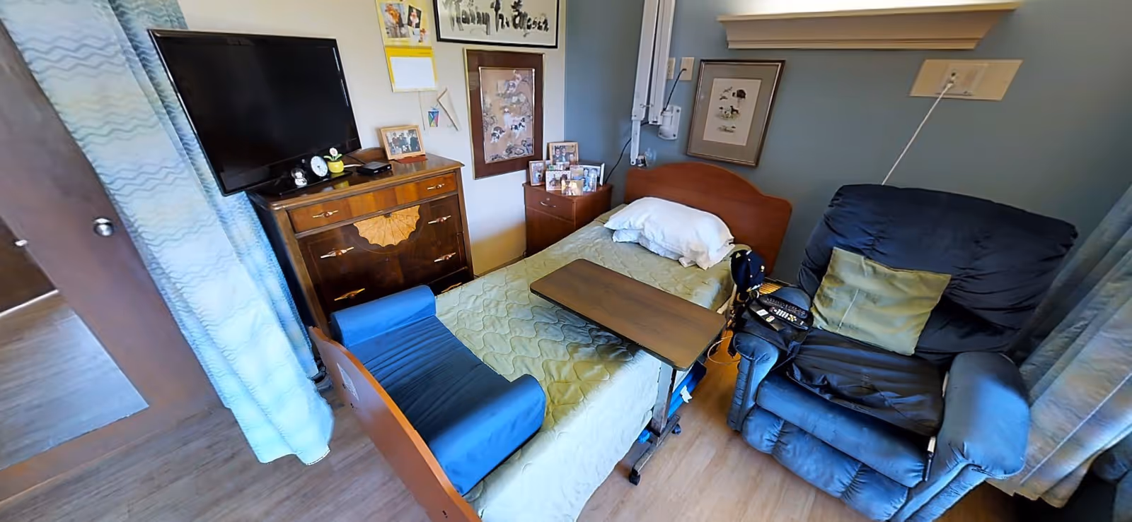 A cozy bedroom with a bed, a wooden dresser, a recliner chair, and a television. The bed is made with a green quilt, and there are personal items and photographs on the dresser.