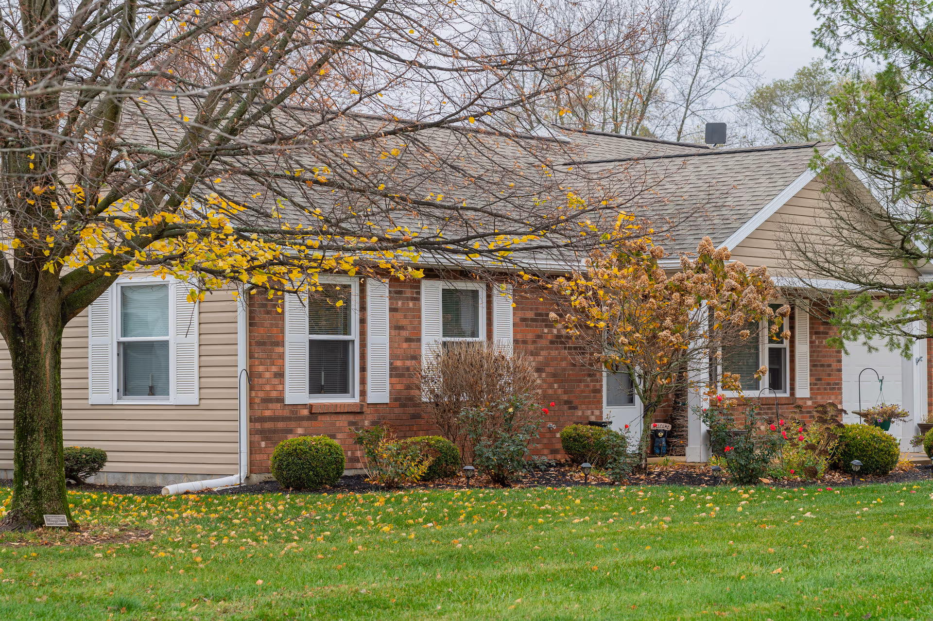Exterior view of a single-story senior living community building with beige siding and red brick walls. The building is surrounded by a green lawn, bushes, and trees with autumn leaves. The sky is overcast.