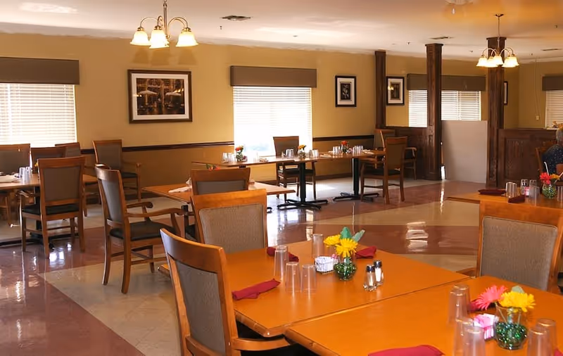 A dining room with several wooden tables and chairs arranged neatly. Each table is set with clear plastic cups, red napkins, salt and pepper shakers, and small vases with colorful flowers. The room has beige walls, windows with blinds, framed pictures on the walls, and ceiling light fixtures providing warm lighting.