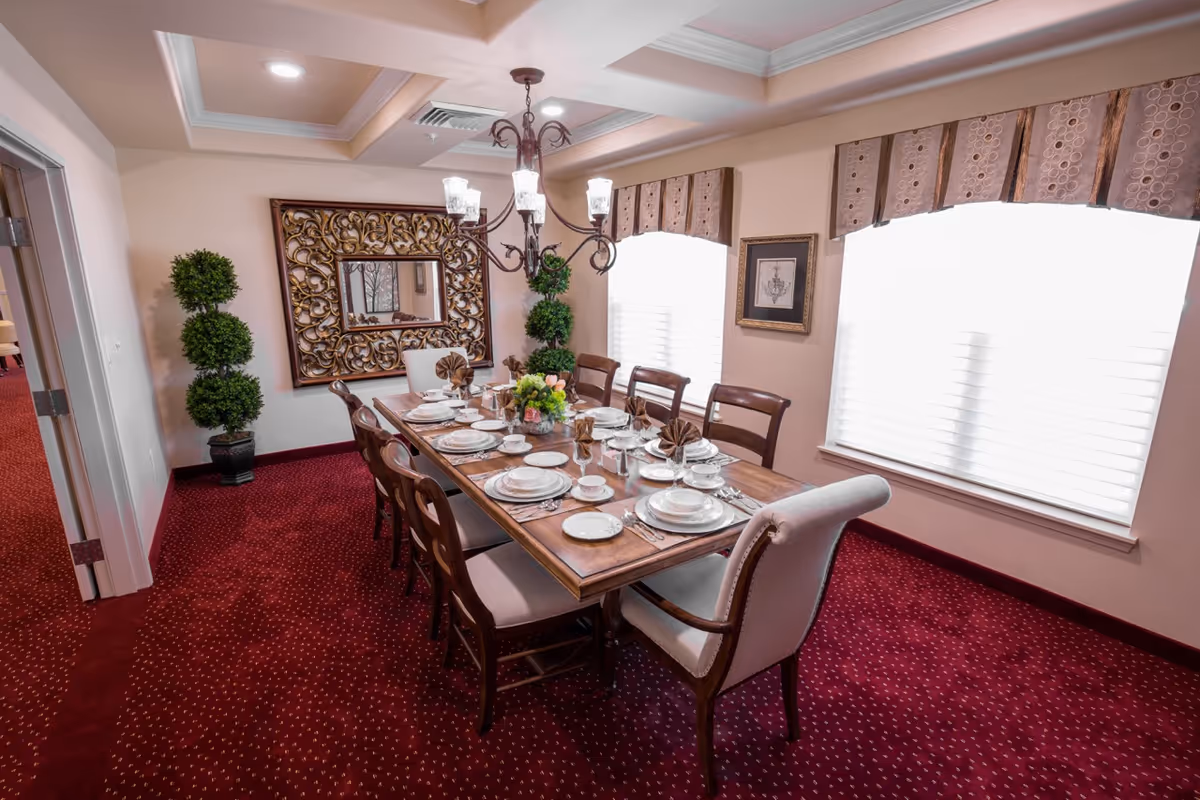 A formal dining room with a long wooden table set for a meal, including plates, cups, silverware, and folded napkins. The room features red carpet with a subtle pattern, beige walls, two large windows with white blinds and decorative valances, a chandelier hanging above the table, a large ornate mirror on one wall, and two potted topiary plants in the corner.