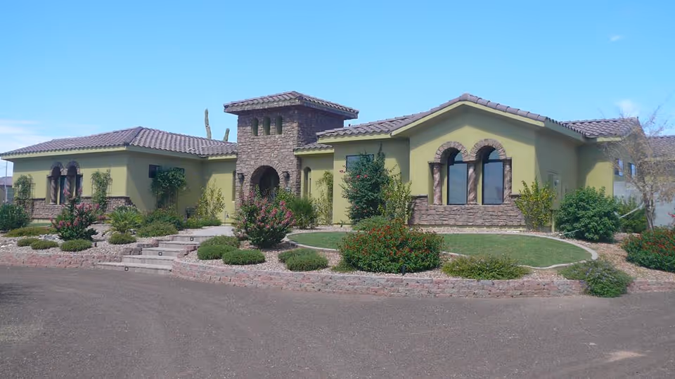 Exterior view of a single-story assisted living facility building with a tiled roof, stone accents around windows and entrance, surrounded by landscaped bushes, plants, and a small lawn under a clear blue sky.