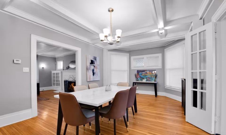 A bright dining room with a rectangular white marble table surrounded by six modern chairs in shades of brown and beige. The room has light gray walls, hardwood floors, and a chandelier hanging from a coffered ceiling. There is a doorway leading to a living room with a fireplace, and a small table with colorful artwork is placed against the far wall beneath three windows with white blinds.