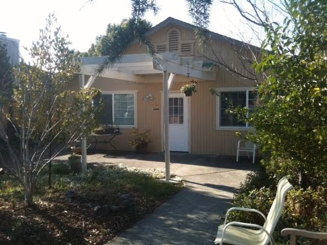 Exterior view of a single-story beige building with a white door and two windows, surrounded by trees and bushes. There is a small covered patio area with a hanging plant and outdoor chairs along a concrete pathway.