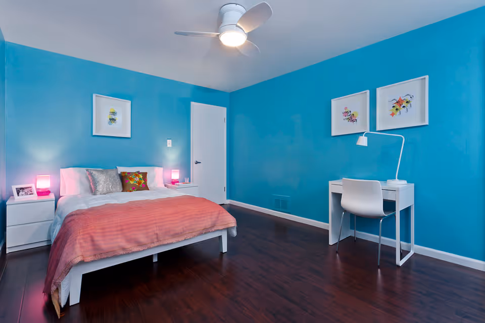 A bedroom with bright blue walls, a white bed with a pink blanket and decorative pillows, two white nightstands with pink lamps, a white door, and a white desk with a chair and a desk lamp. Three framed artworks hang on the walls.