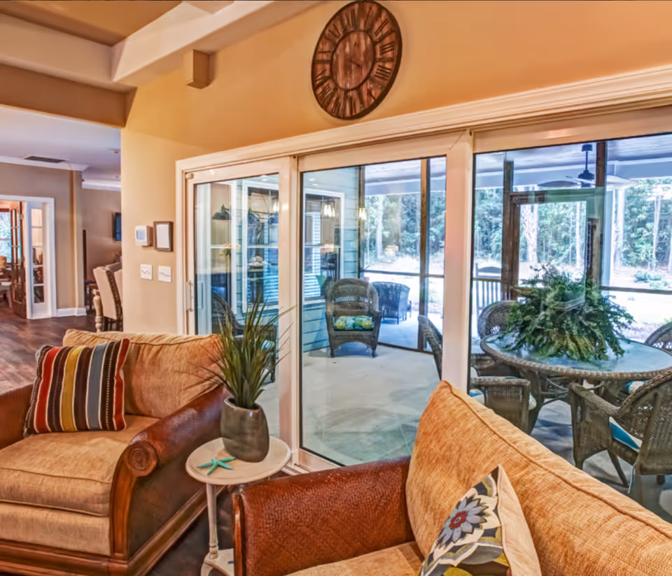 Cozy living room area with two cushioned armchairs featuring patterned pillows, a small round side table with a potted plant and a decorative starfish, large sliding glass doors leading to a screened outdoor patio with wicker chairs and a round table with a green plant centerpiece, and a large wall clock above the doors.