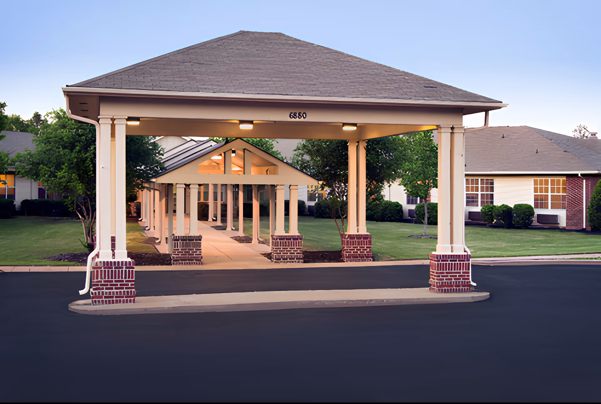 Covered entrance with a peaked roof supported by columns with brick bases, leading to a walkway lined with similar columns and surrounded by green lawns and trees. Residential buildings with large windows are visible in the background under a clear sky.