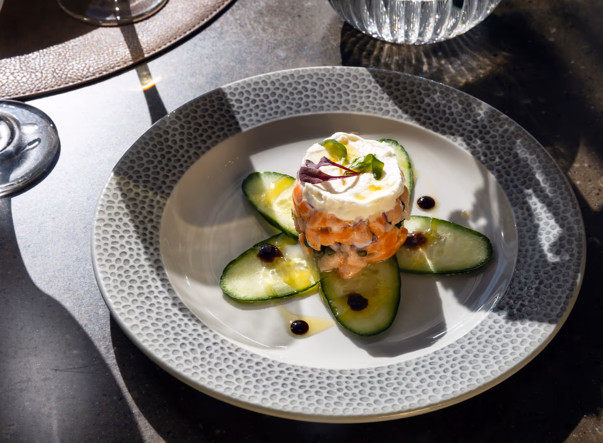 A white plate with a textured gray rim holding a gourmet appetizer consisting of a circular stack of diced salmon topped with a dollop of cream and garnished with small greens, surrounded by cucumber slices and small drops of dark sauce, placed on a table with a glass and a water pitcher nearby.