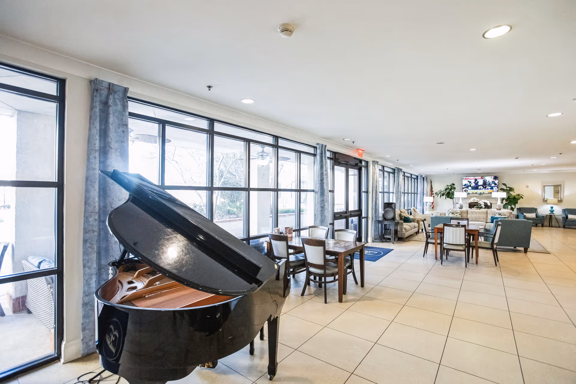 A spacious and bright common area with large windows along one side, featuring a black grand piano in the foreground, several tables and chairs, and comfortable seating areas with sofas and armchairs. A television is mounted on the far wall above a fireplace, and there are plants and decorative items around the room.