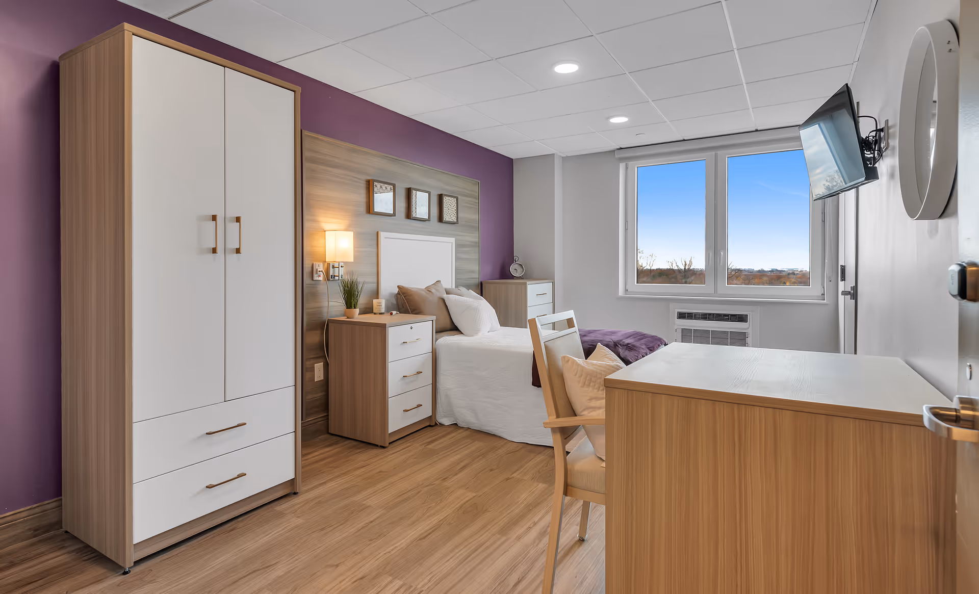 A bright and clean assisted living bedroom with a single bed, wooden wardrobe, nightstand with lamp, dresser, desk with chair, wall-mounted TV, and a large window showing a clear blue sky outside. The room features a purple accent wall and wood flooring.