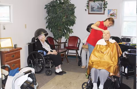 A senior living facility room where an elderly woman in a wheelchair is getting her hair styled by a caregiver. Another elderly woman in a wheelchair is sitting under a hair dryer. The room has chairs, a potted plant, framed pictures on the wall, and windows letting in natural light.