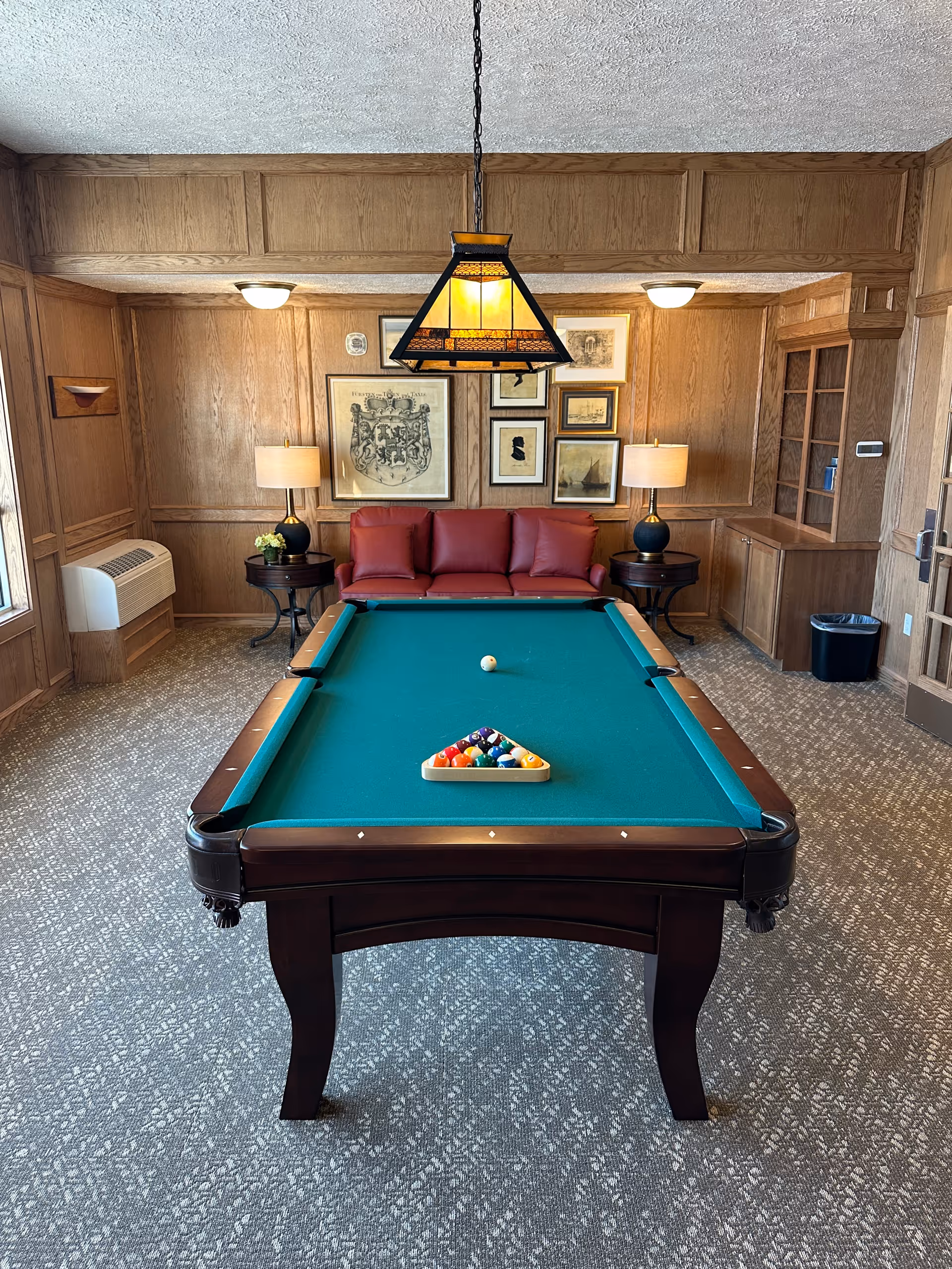 A cozy game room with a green felt pool table in the center, a red leather couch against a wood-paneled wall, two side tables with lamps, framed artwork on the wall, and a hanging light fixture above the pool table.