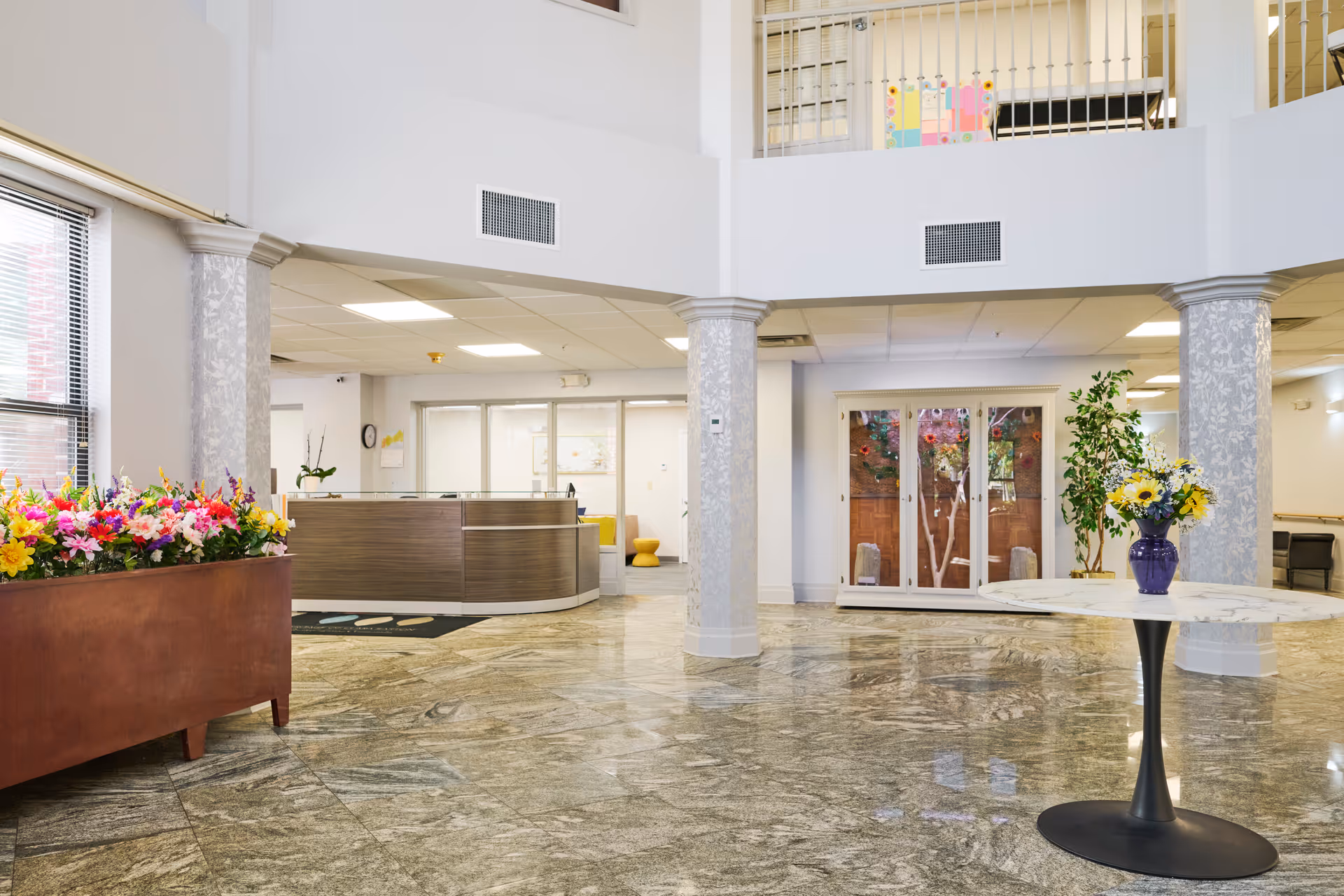 Spacious senior living facility lobby with a reception desk, marble floor, decorative columns, and flower arrangements.
