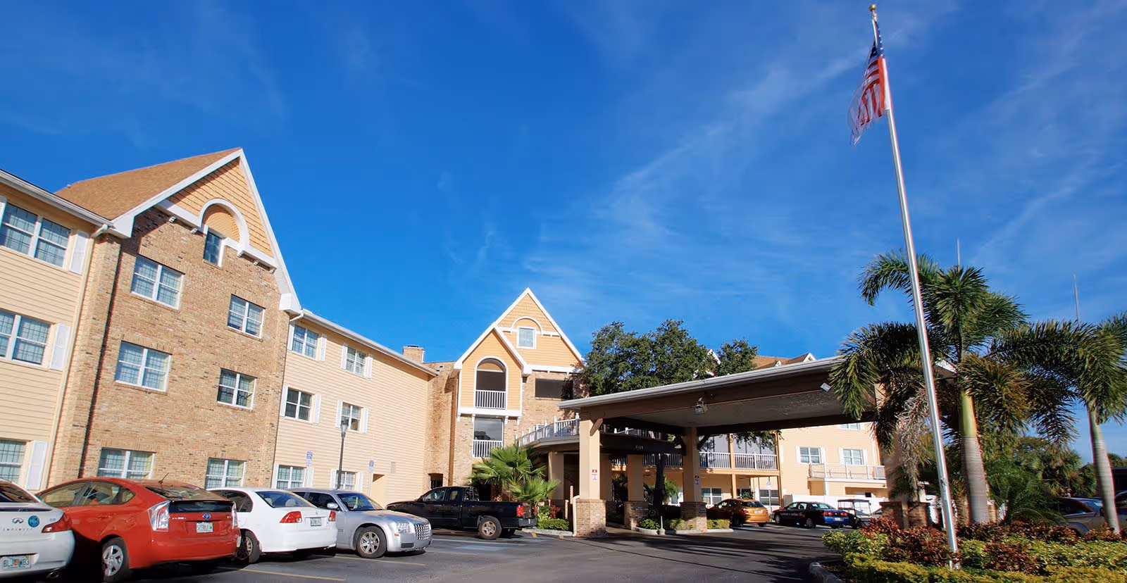Exterior view of Bayside Terrace senior living facility showing a multi-story building with beige siding and brick accents, a covered entrance, several parked cars, palm trees, and an American flag on a flagpole under a clear blue sky.