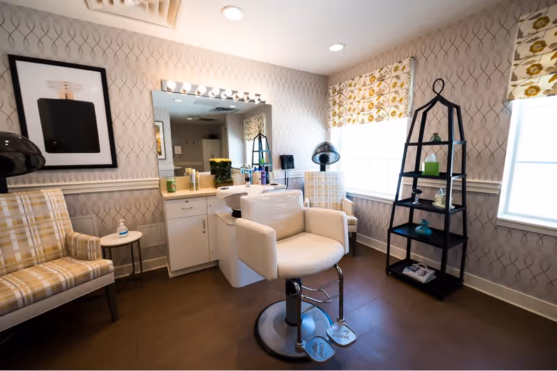 Well-lit salon/styling room with a white styling chair in front of a mirror, hooded dryers, plaid armchairs, and a black shelving unit by two windows.