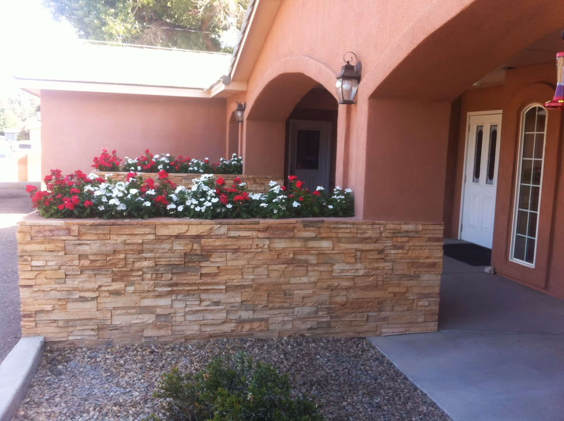 Exterior view of a building entrance with a stone planter filled with red and white flowers. The building has a peach-colored stucco finish with arched doorways and white double doors. There are outdoor wall lanterns mounted on the walls near the entrance.