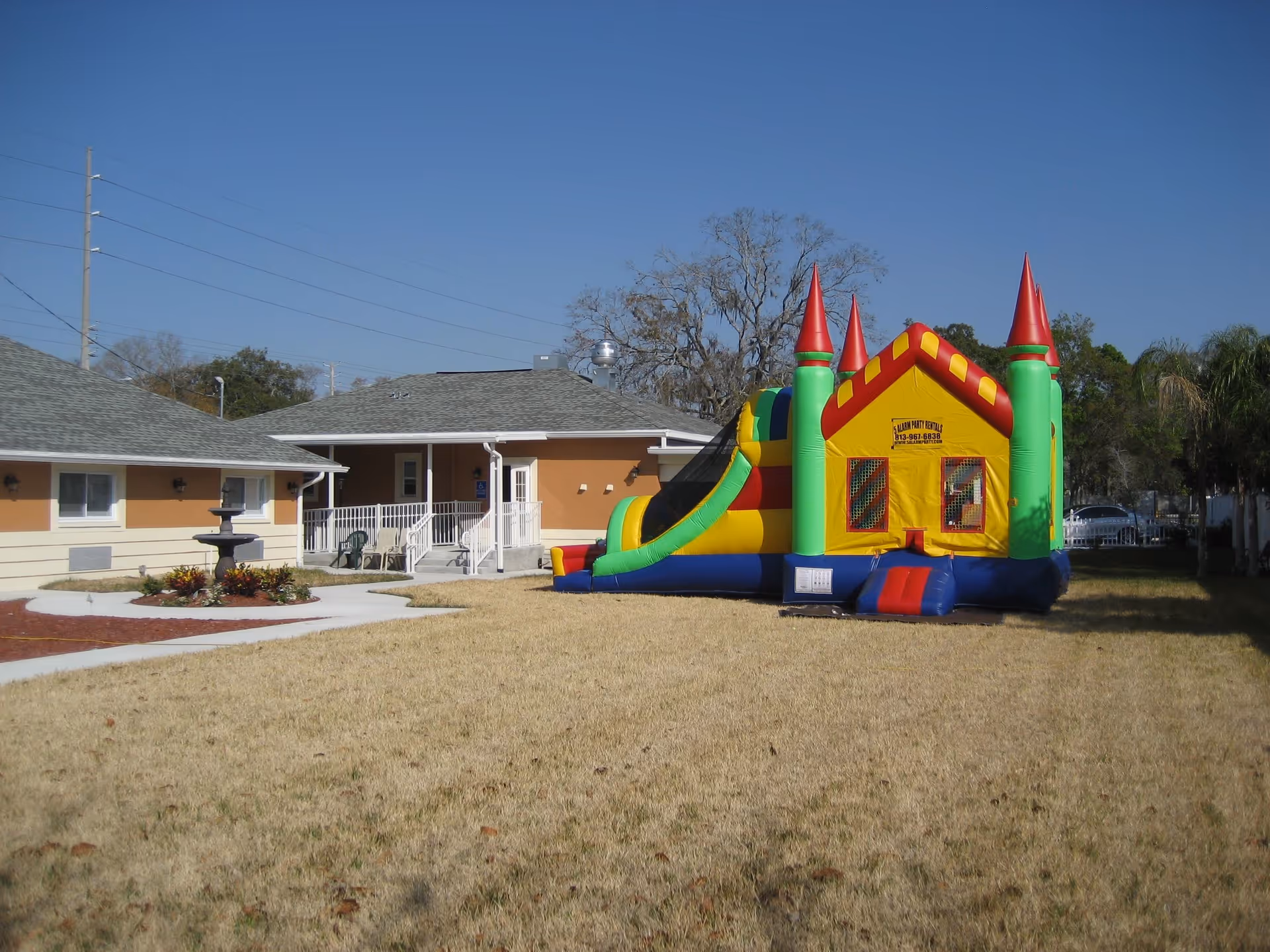 Outdoor area of Bamboo Villas Alf facility featuring a large inflatable bounce house with slide on a grassy lawn, adjacent to a beige building with a gray roof and a small fountain surrounded by plants.