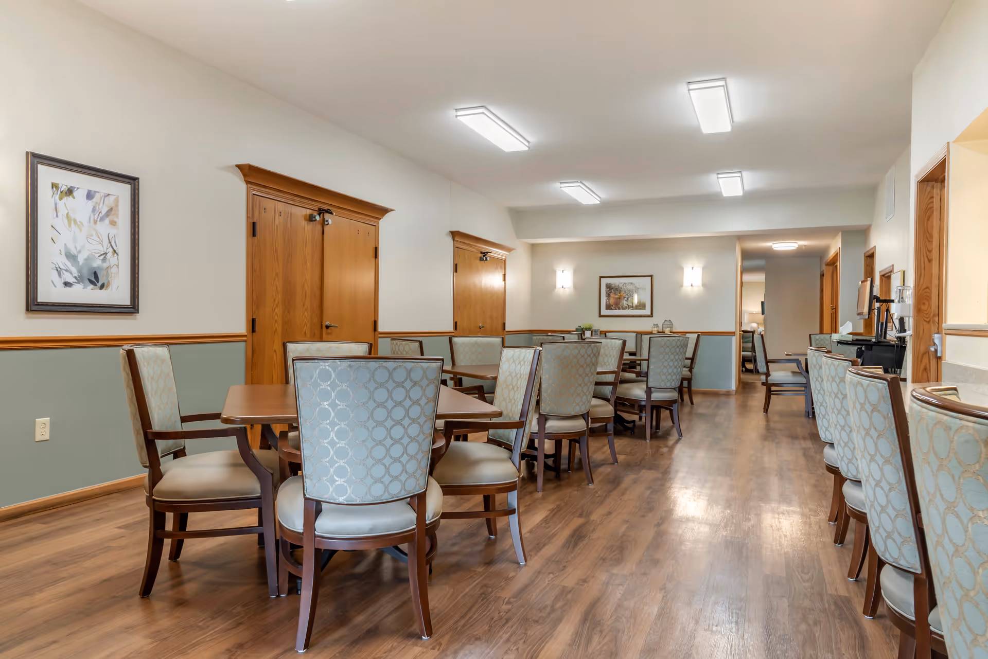 A bright communal dining room with multiple wood tables and upholstered chairs on a hardwood floor.