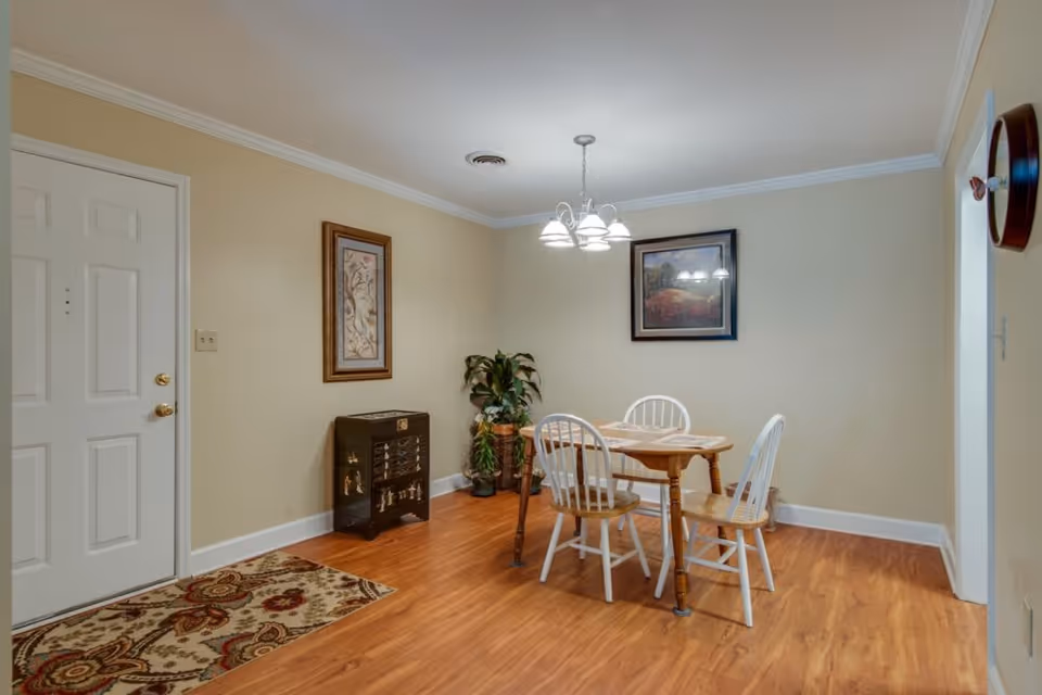 A dining area with a wooden table and four chairs, three of which are white and one is wooden. The room has light beige walls, a wooden floor, a hanging light fixture above the table, two framed pictures on the walls, a small decorative cabinet, a potted plant, and a patterned rug near a white door.
