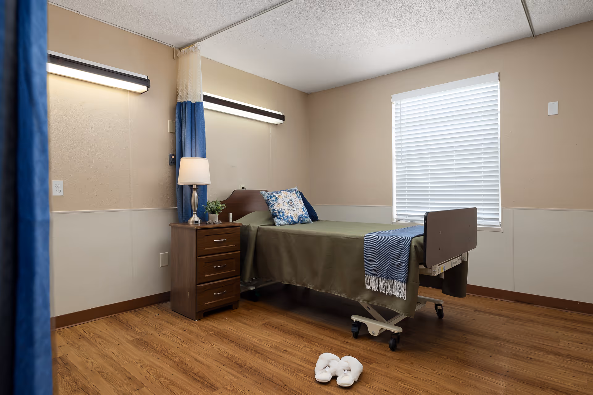 A simple nursing facility bedroom with a single bed covered in an olive green blanket and a blue throw. There is a wooden nightstand with a lamp and a small plant next to the bed. The room has beige walls, a window with white blinds, and wooden flooring. A pair of white slippers is placed on the floor near the foot of the bed.