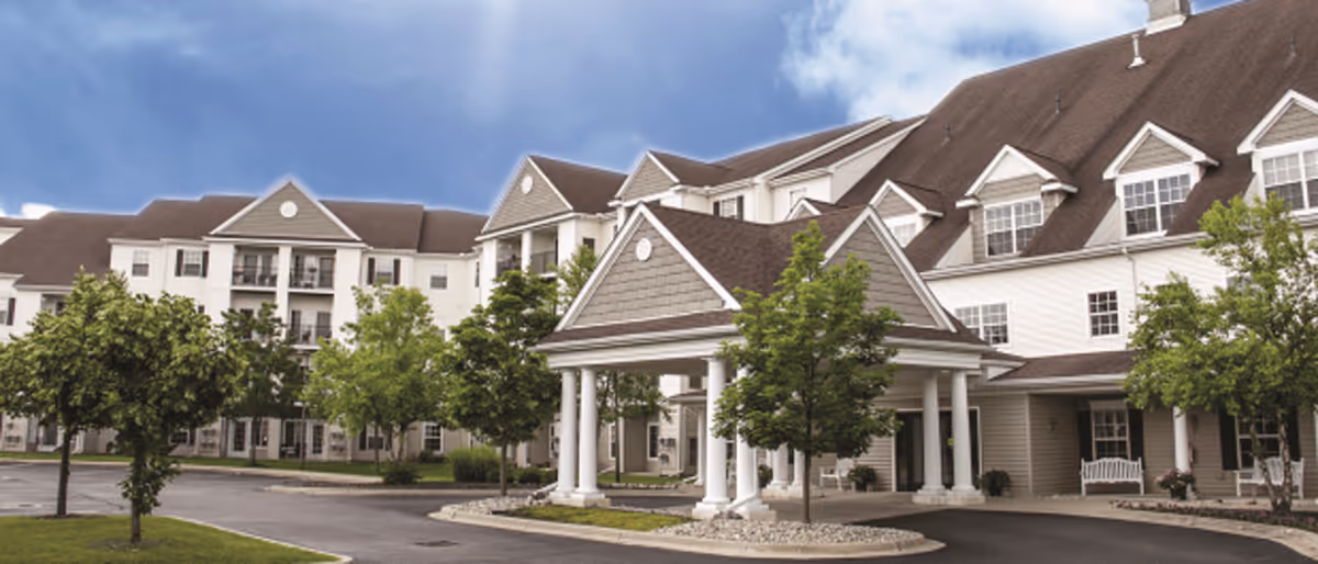 Exterior view of a large senior living facility building with multiple floors, a covered entrance supported by white columns, several trees, and a paved driveway under a partly cloudy sky.