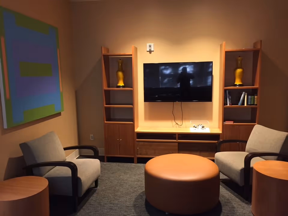 A cozy living room area with two gray armchairs with dark wooden armrests, a round brown ottoman in the center, and two wooden shelving units on either side of a wall-mounted flat-screen TV. The shelves contain yellow vases and some books. There is a colorful abstract painting on the left wall and a small round wooden side table next to one of the armchairs.