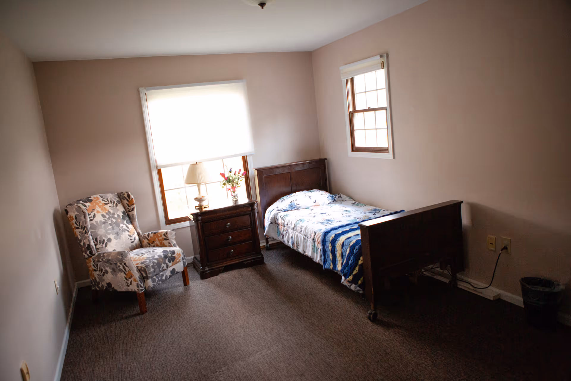A small bedroom with a single wooden bed covered with a floral and striped blanket, a wooden nightstand with a lamp and a vase of flowers, a floral upholstered armchair, two windows with white blinds, beige walls, and brown carpeted floor.