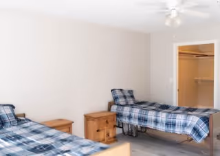 A bedroom with two single beds covered in blue plaid bedding, a wooden nightstand between them, and an open door revealing a closet with shelves and hanging space.