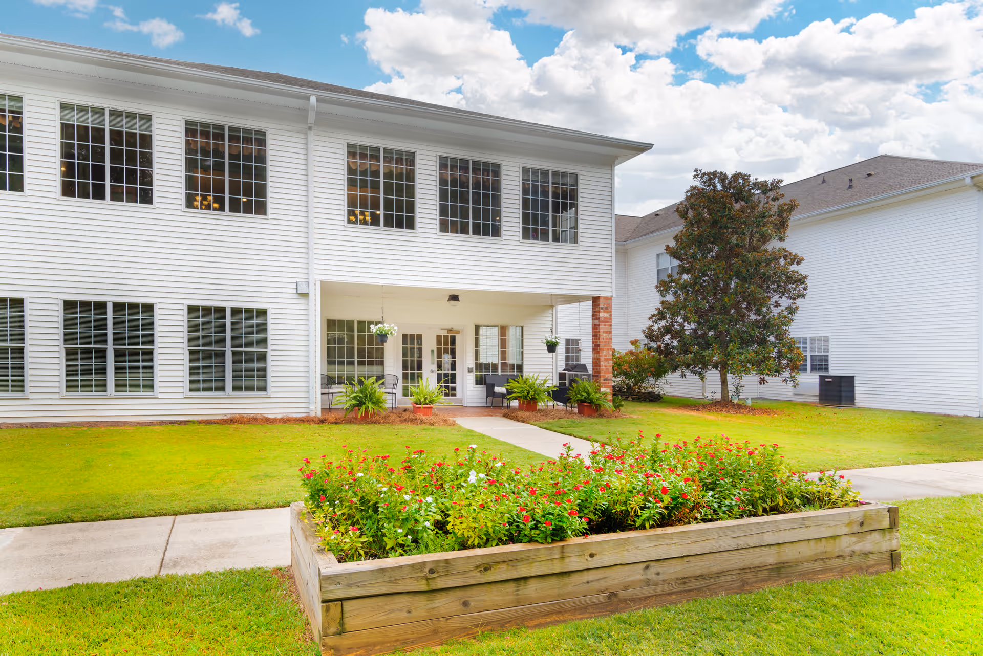 Exterior view of a two-story white building with large windows, a covered patio area with chairs and hanging plants, a flower bed with red and white flowers in the foreground, and a tree on the right side under a partly cloudy sky.