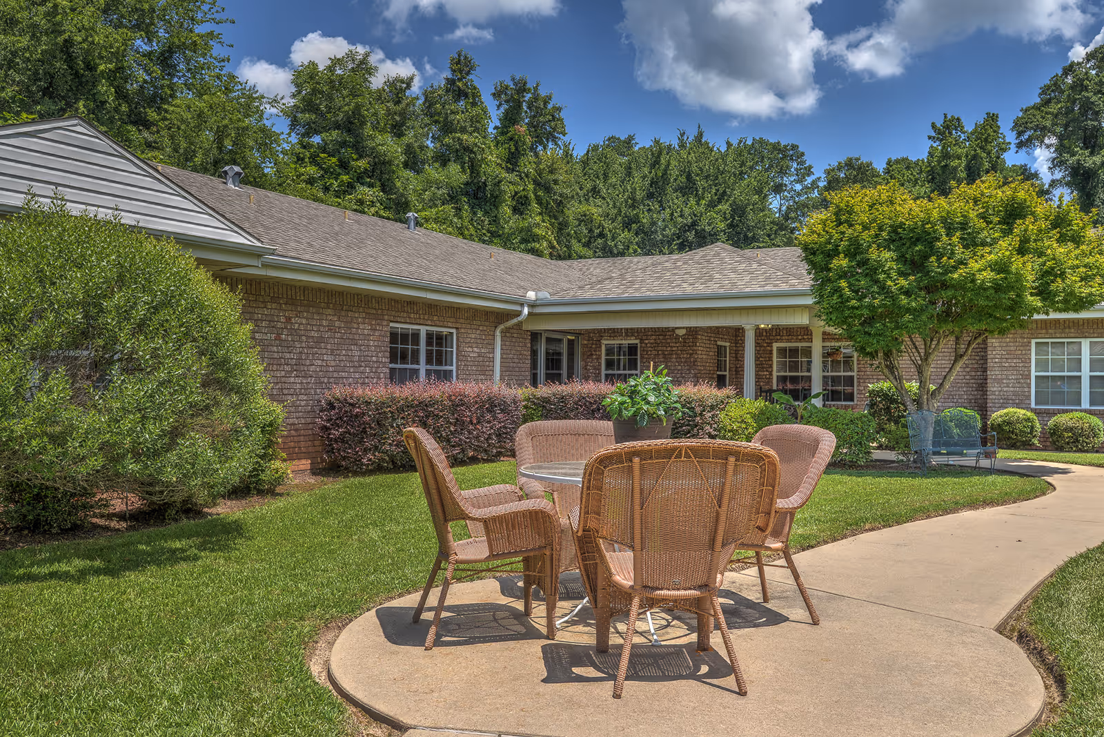 Outdoor patio area at Oakwood House featuring a round glass table with four wicker chairs on a concrete pathway surrounded by green grass, bushes, and trees with a brick building in the background under a partly cloudy blue sky.